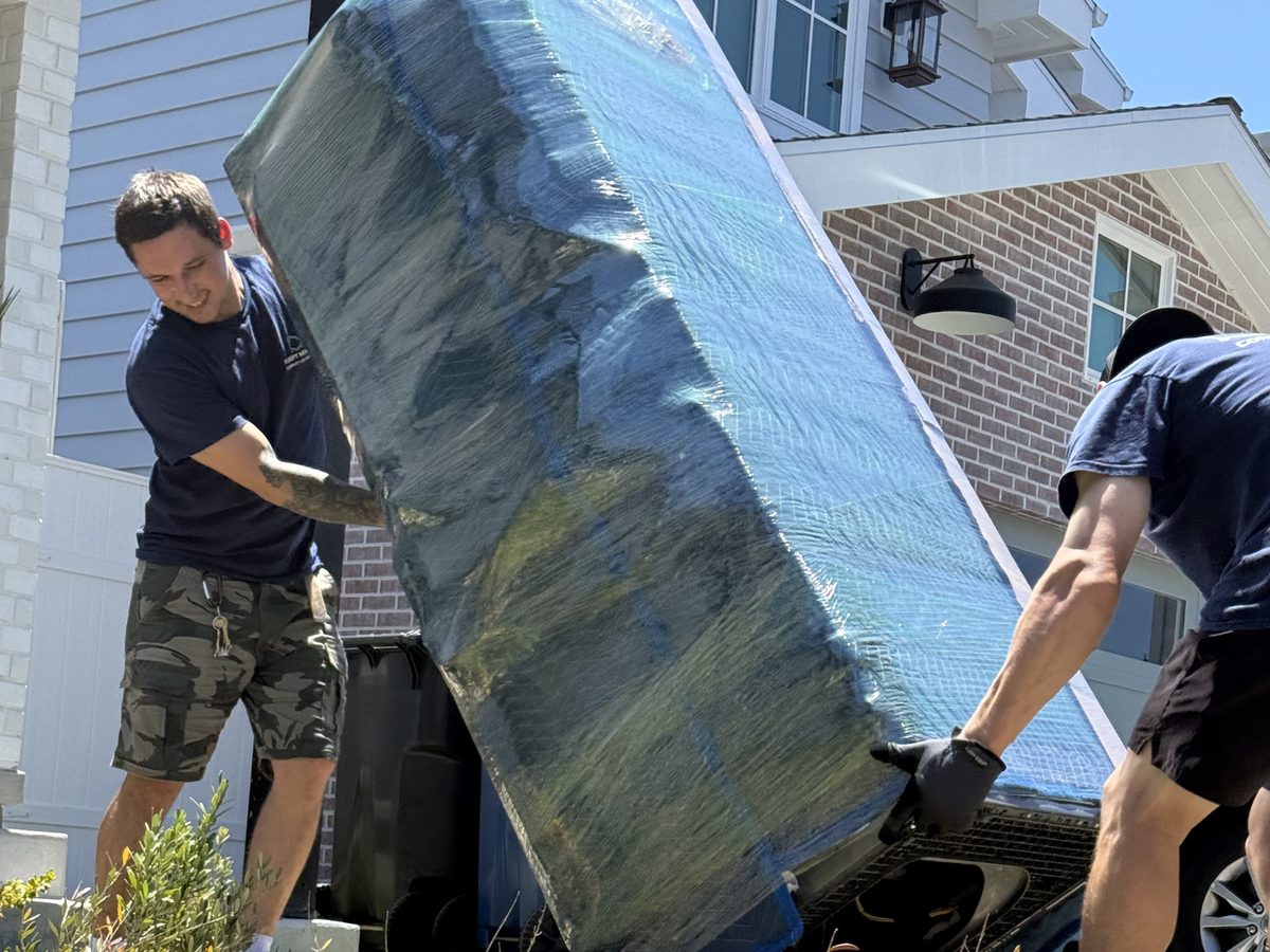 Two Adept Moving movers rolling a blanket-wrapped fridge across the front lawn on Wendy Way