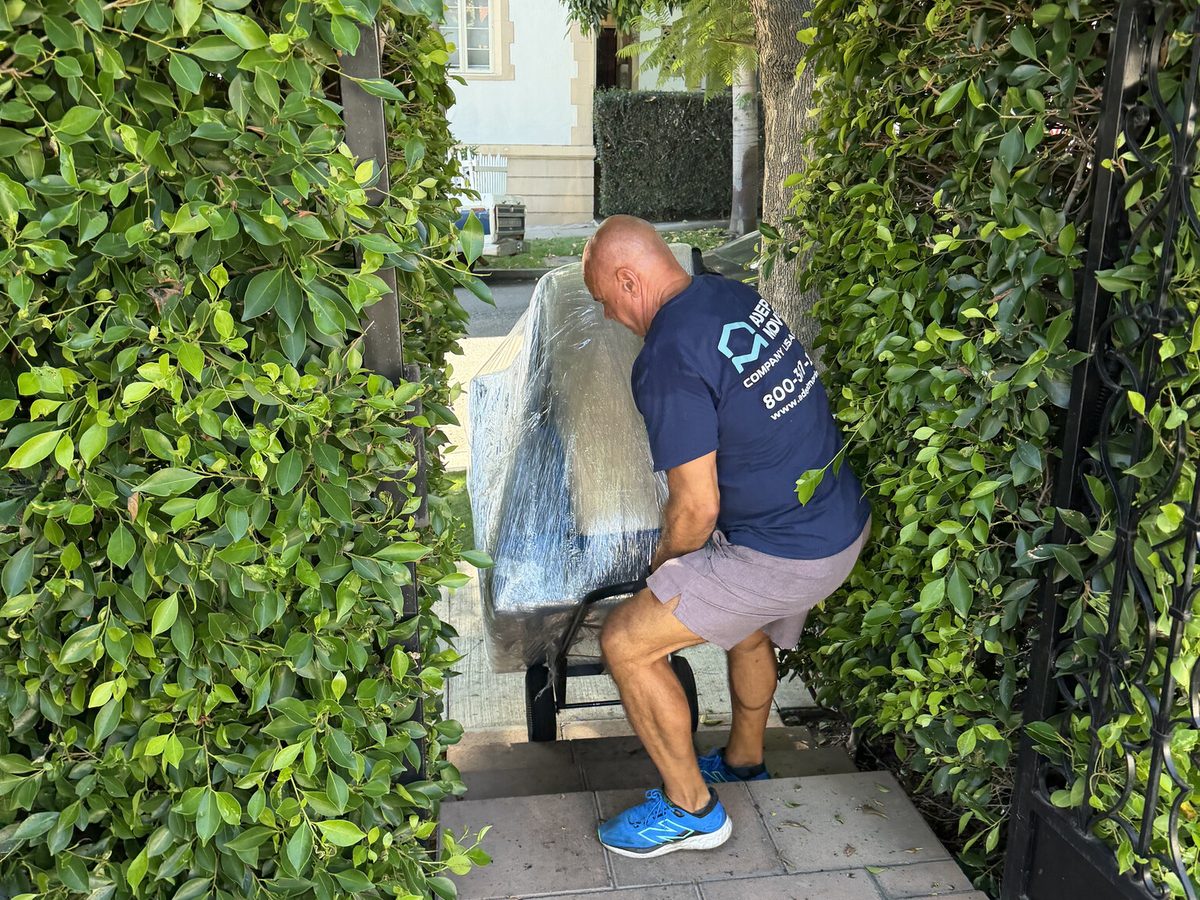 Adept Moving mover carrying a blanket-wrapped piece along the hedged walkway of a West Hollywood courtyard building