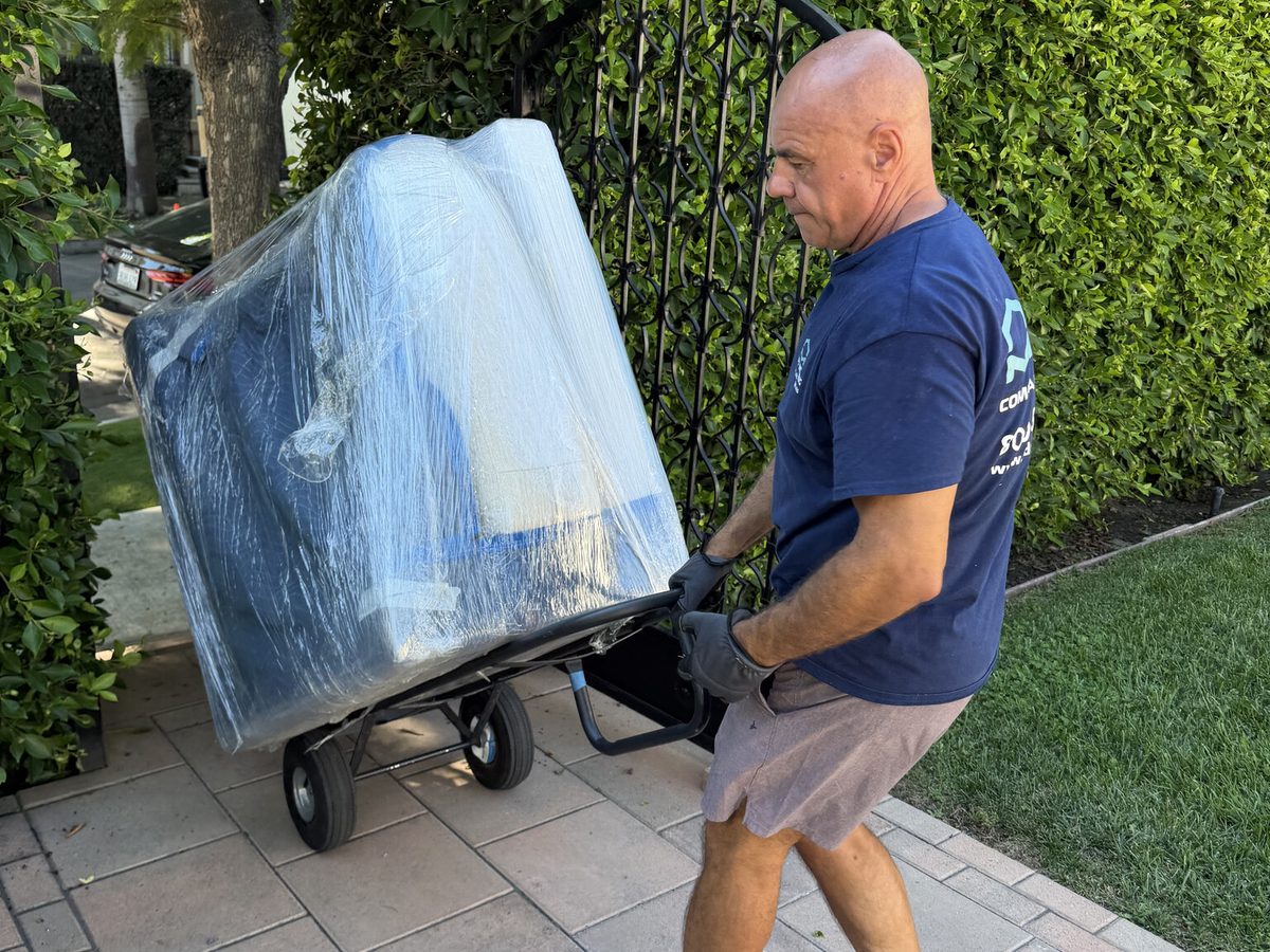 Adept Moving mover turning a wrapped sofabed on a hand truck at the wrought-iron gate on North Harper Avenue