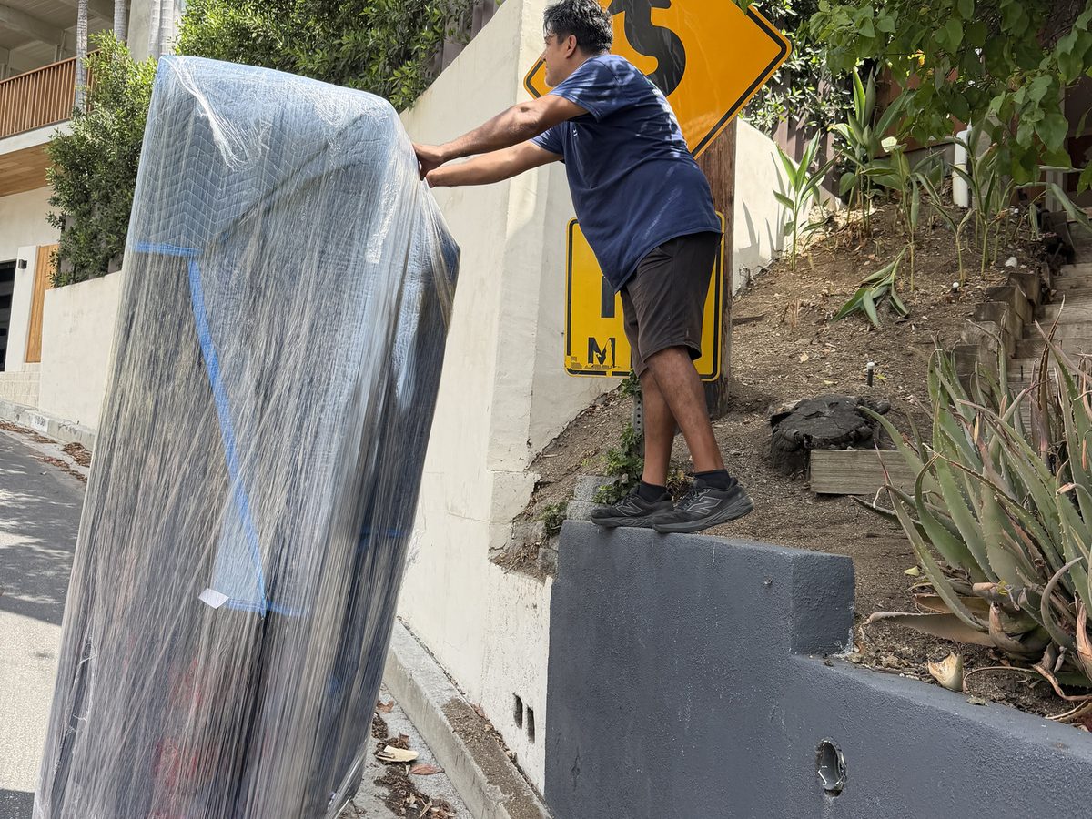Adept Moving mover walking a shrink-wrapped mattress up past the retaining wall on the hillside street
