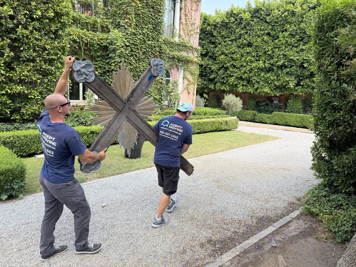 Two Adept Moving movers carrying the wooden cross with a gilded sunburst along the driveway