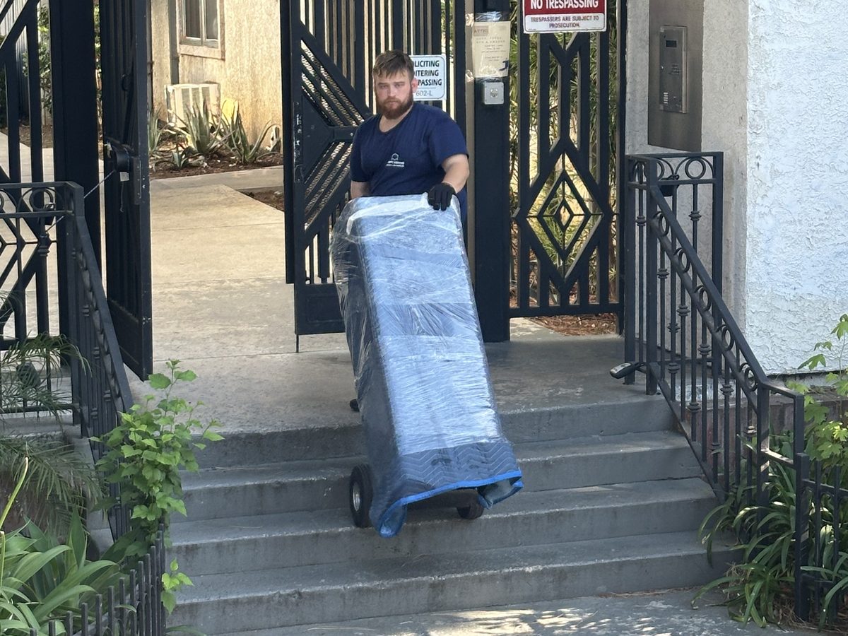 Adept Moving mover wheeling a wrapped piece on a hand truck down the steps at the gate of the condominium complex